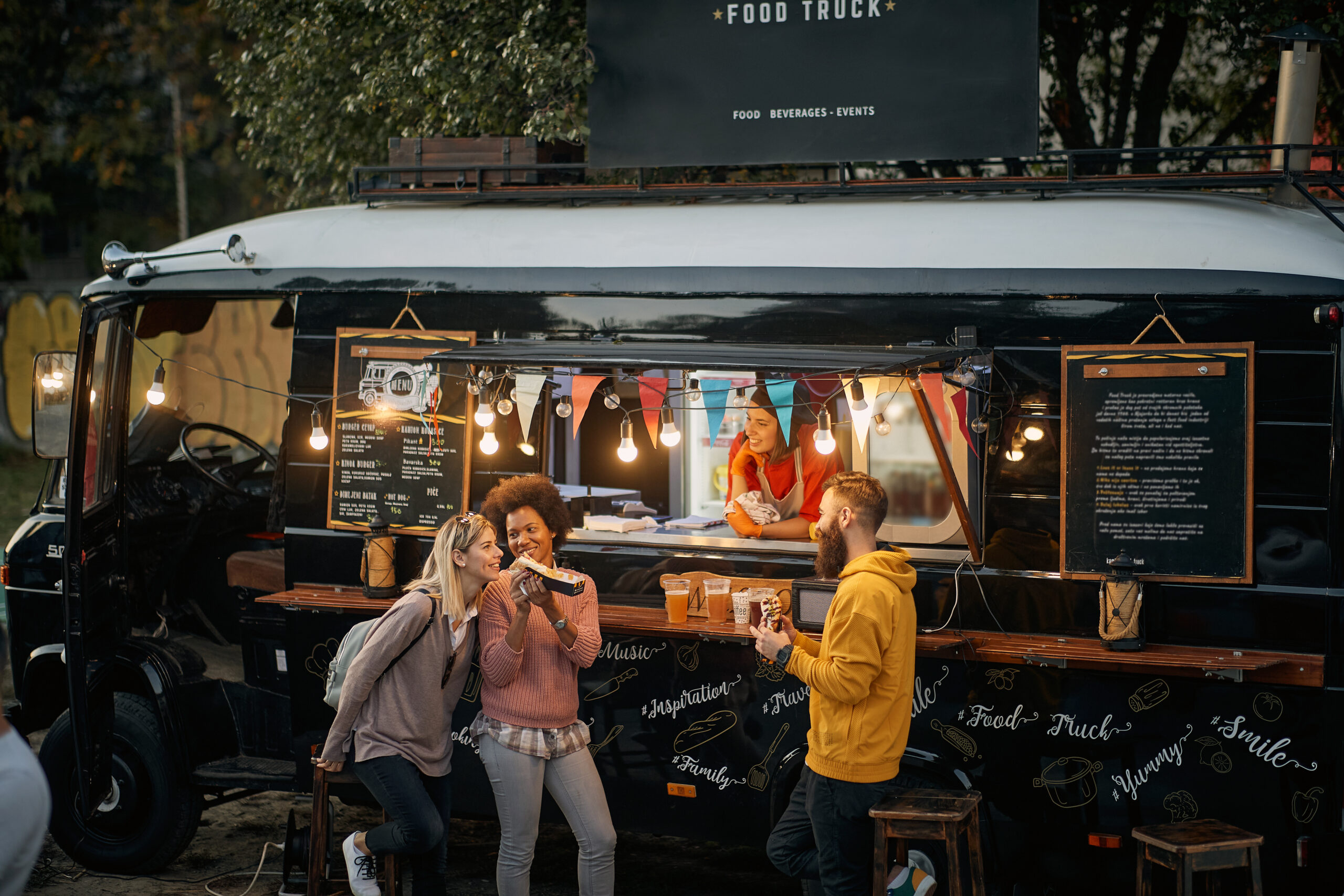 A multiethnic group of young people socializing while eating outdoor in front of modified truck for fast food.